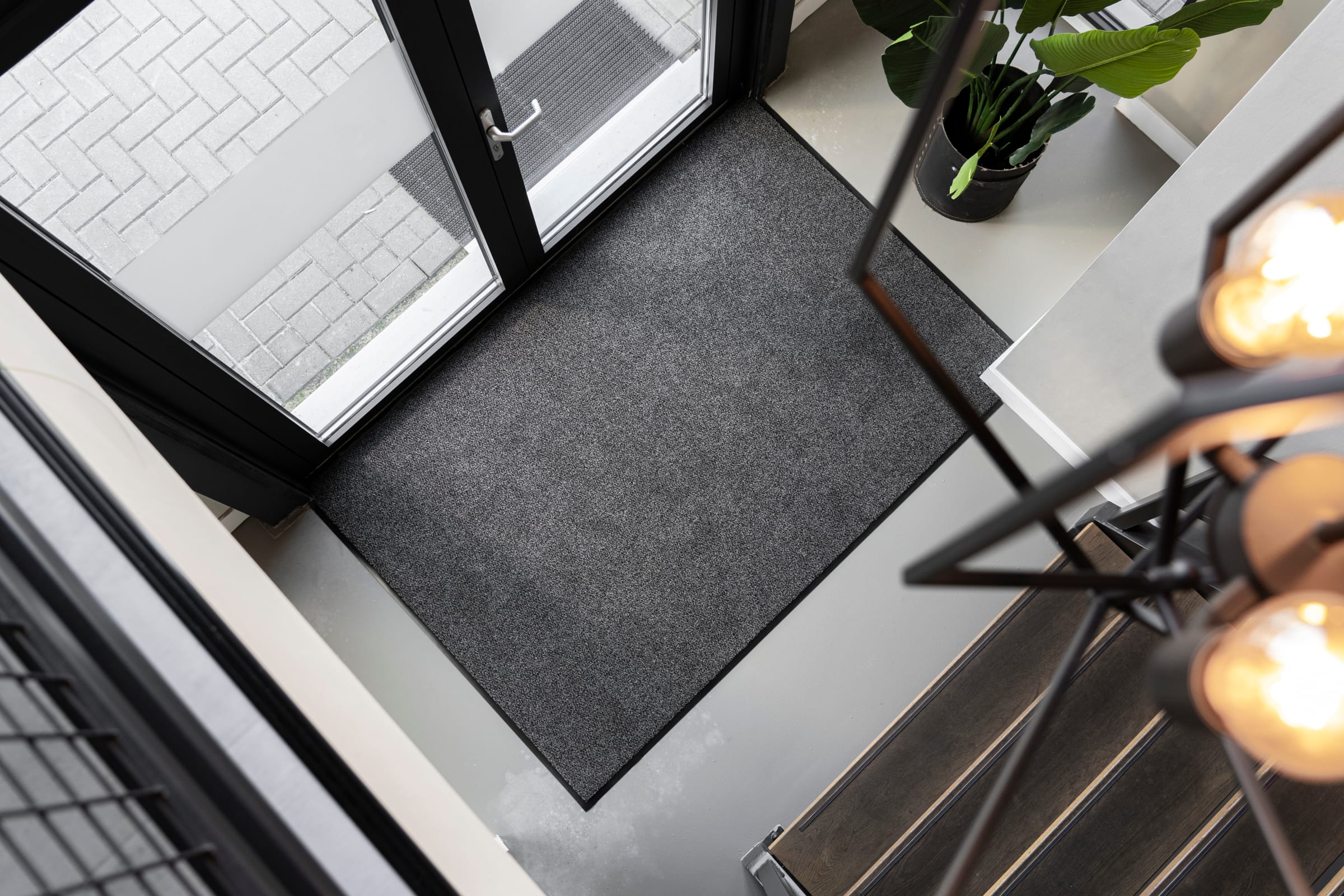Praktijkvoorbeeld van de Ingresso Schoonloopmat. De mat is hier te zien in een thuissituatie, waarbij ook een plant, lamp en voordeur op de foto staan.